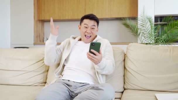 兴奋的亚洲年轻人 拿着手机坐在沙发上 欢呼雀跃地赢了体育赌局 好消息 快乐的年轻人用手机庆祝网上拍卖的胜利 收到了阅读短信 — 图库视频影像