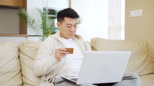 ラップトップコンピューターとクレジットカードを使ってインターネットストアで買い物をしている若い幸せなアジア人男性 男性は自宅のリビングルームのソファに座っているアプリケーションでのサービスやチケットの支払いデータを入力します — ストック動画