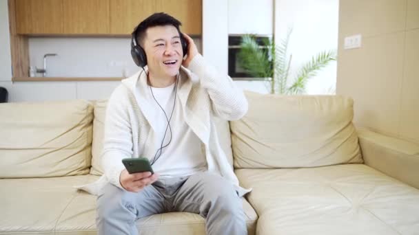 自宅のソファに座ってヘッドフォンで音楽を聴いている若い陽気なアジアの男 ソファの上の男だけでいい生活を感じている 電話で幸せな男性はオンラインで彼のお気に入りのラジオ局を聞く — ストック動画