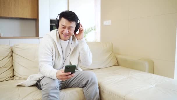 Young Cheerful Asian Guy Listening Music Headphones Sitting Sofa Home — Stock Video