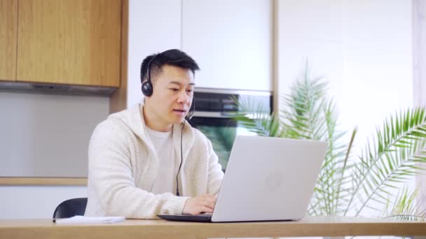 年轻的亚洲人自由职业者坐在家里的写字台前 带着耳机 通过笔记本电脑在房间或厨房里上网聊天 穿着休闲装的男人在屋里干活 — 图库视频影像