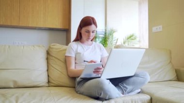Öğrenci kız evde online çalışıyor. - İnternet. Laptop video dersleriyle birlikte e-öğrenme. Kanepede oturan genç bir kadın not defterine not tutuyor ve ders veriyor. Uzaktan eğitim. Öğrenim evi kanepede.