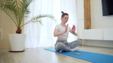 Genç ve çekici bir kadın internette cep telefonu ya da akıllı telefon uygulaması kullanarak yoga yapıyor. Kadın yogi sabahı evde günlük kıyafetlerle oturma odasında, lotus pozisyonu bir minder üzerinde meditasyon yapar..
