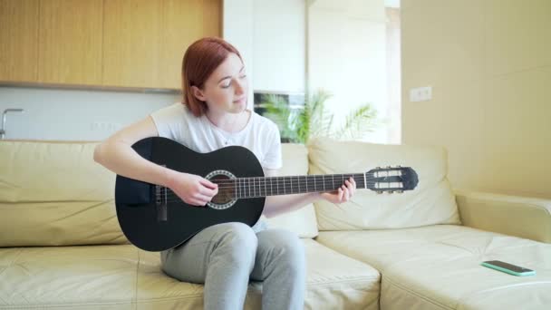 Młoda Ruda Dziewczyna Siedzi Kanapie Gra Gitarze Akustycznej Biała Studentka — Wideo stockowe
