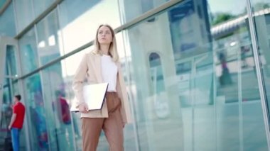 Genç bir iş kadını modern bir ofis binasının arka planında şehir caddesinde yürüyor. İş kadınıyım. Kendine güveni tam beyaz bir kadın elinde dizüstü bilgisayarla dışarı çıkıyor. 
