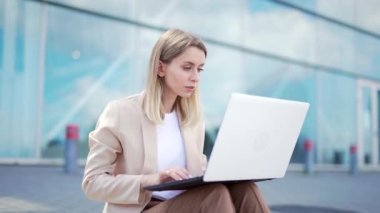 Genç iş kadını kaldırımda oturuyor ve modern ofis merkezinin dizüstü bilgisayarında çalışıyor. İş kadını, kız öğrenci üniversitesi ya da açık havada üniversite. Şehir caddesinde