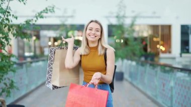 Hediyelik poşetlerde alışveriş yaparken alışveriş merkezinde kameraya bakan mutlu, genç bir kadın portresi. Neşeli kadın alıcı sevinir ve satış pozu verir. Kara Cuma. Paket tutan güzel kız.