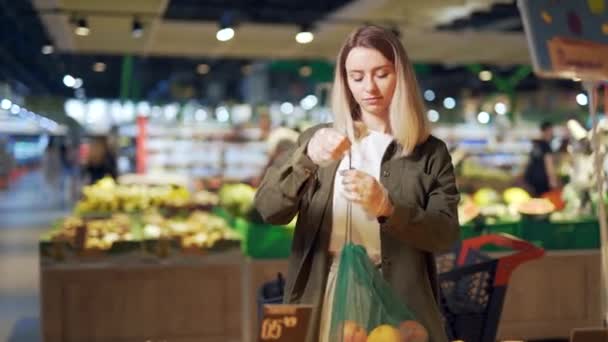 若い女性はエコバッグ野菜や果物を選ぶスーパーマーケットのオレンジ カウンター近くの食料品店に立つ女性客は市場で再利用可能なパッケージを購入し投げます — ストック動画