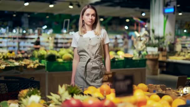 野菜コーナーのスーパーに立つ腕の中でポートレート若い本格的な自信女性労働者が交差した 緑の果物店の市場でカメラを見て売り手の女性 作業エプロンの従業員の八百屋 — ストック動画