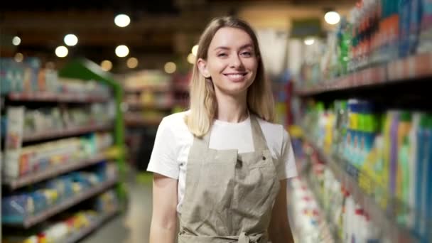 女售货员的肖像 女售货员笑着看着超市里的相机 快乐友善的女店员站在商店的两排中间 商业和人员概念 — 图库视频影像