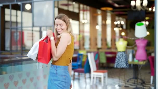 モールに立っている間にカメラを見てギフトの紙袋にショッピングを示す肖像幸せな若い陽気な女性 喜びの女性のバイヤー喜びとポーズ販売 ブラック フライデー かわいい女の子がパケットを保持 — ストック動画