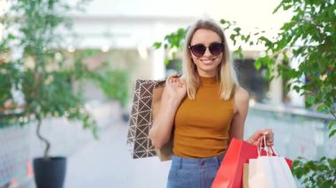 Hediyelik poşetlerde alışveriş yaparken alışveriş merkezinde kameraya bakan mutlu, genç bir kadın portresi. Neşeli kadın alıcı sevinir ve satış pozu verir. Kara Cuma. Paket tutan güzel kız.