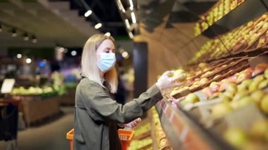 Koruyucu maskeli genç sarışın kadın süpermarkette elma meyvesi seçiyor. Markette kadın alışverişi. Alışveriş merkezinin yanında elinde sepetle duruyor.