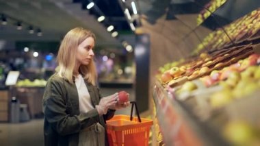 Genç sarışın kadın süpermarkette sebze topluyor. Markette alışveriş yapan kadın elinde sepetle sebze mağazasının yanında duruyor. Elmayı inceler