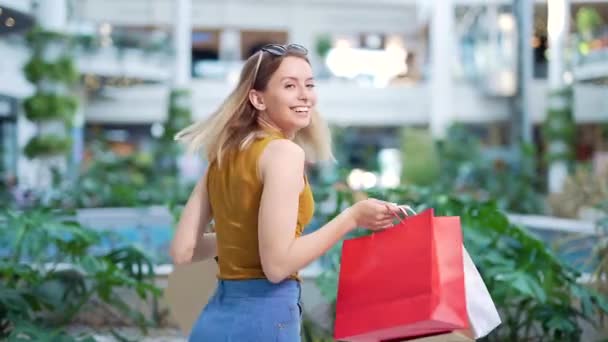 買い物袋を持っている陽気な若いお店の女性は ショッピングセンターで踊るカメラを見て笑っています 幸せな女性持っている楽しいです黒金曜日楽しくジャンプのために喜びでザ モール — ストック動画