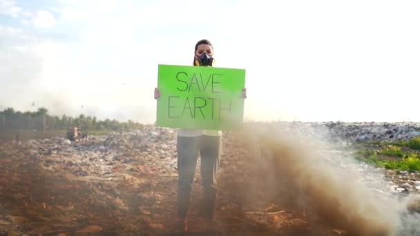 手拿着海报拯救地球的年轻女权运动者站在一个有呼吸器脸的垃圾填埋场中间 女生抗议环境污染及自然保育 — 图库视频影像