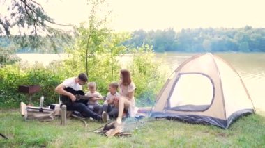 Ormanda göl kenarında kamp yapmak. Mutlu aile babası, anne ve küçük çocuklar, doğada ateş ve çadır başında oturuyorlar. Tatilde birlikte vakit geçirin. Açık havada. Çocuklu ebeveynler. Babam gitar çalıyor. Kamp