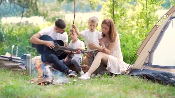 森の中の湖でキャンプ 幸せな家族のお母さんと小さな子供たちが自然に火とテントに座っています 余暇を一緒に休暇で過ごしましょう 屋外だ 子供を持つ親 父はギターを弾いてた キャンプ — ストック動画