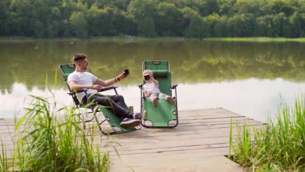 小さな息子と幸せな父親は 湖の木製の桟橋に快適な椅子に座って休んでいる 父と子供の男の子は屋外で一緒に余暇を過ごします 田舎でのキャンプ休暇 父の日 — ストック動画