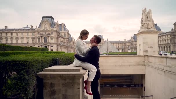 ルーブル美術館を背景にパリの屋外で一緒にキスを愛するスタイリッシュな若いカップル ヨーロッパで歴史的に美しい場所 幸せと愛のボーイフレンドとガールフレンドで ロマンチックなデートと会議 — ストック動画
