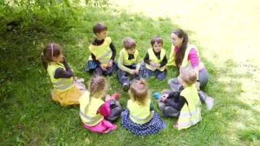 Bir grup küçük çocuk bir öğretmenle bir çemberde oturuyor ve parkta ya da ormanda birlikte oyun oynuyorlar. Anaokulu gezisi ya da yürüyüş. Çocuklar çim çayırında oturup eğleniyorlar. Yaz kampı açık havada