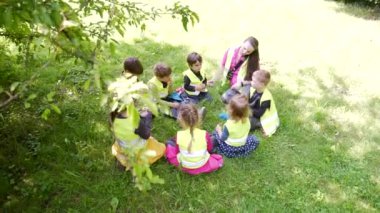 Bir grup küçük çocuk bir öğretmenle bir çemberde oturuyor ve parkta ya da ormanda birlikte oyun oynuyorlar. Anaokulu gezisi ya da yürüyüş. Çocuklar çim çayırında oturup eğleniyorlar. Yaz kampı açık havada