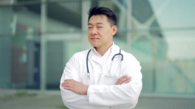 Dışarıdaki modern hastanenin arka planında kollarını kavuşturmuş kameraya bakan Asyalı doktorun portresi. Kendine güvenen bir doktor. Erkek bilim adamı terapist kliniğin yakınında önlük giyiyor.