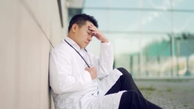 Stresli bir Asyalı doktorun portresi bir hastanenin önünde ya da dışında duruyor. Kliniğin yakınında düşünceli bir doktor. Salgın sırasında steteskoplu sağlık görevlisinin stresi.