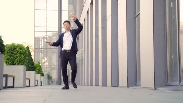 陽気で幸せなアジアのビジネスマンは モダンなオフィスビルを背景に廊下を歩いて踊る 屋外スーツで面白い成功したビジネスマンは喜んでジャンプは勝利を祝う — ストック動画