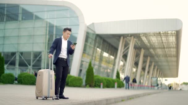 一名亚洲商人站在一个身穿正装的现代化火车站机场的后面 他带着一个手提箱 用的是应用手机 随手拿着智能手机旅游的商人 — 图库视频影像