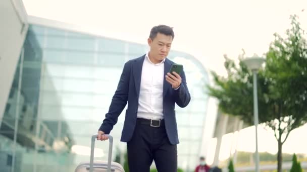 アプリの携帯電話を使用してスーツケースとの正式なスーツケースで近代的な鉄道駅の空港を背景に立ってアジアのビジネスマン スマートフォンを手にビジネスマンの観光客を旅行スペースをコピー — ストック動画