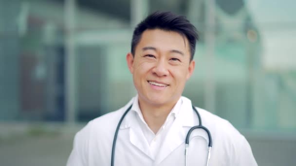 外の近代病院を背景に腕を組んでカメラを見ているアジア人医師の肖像画を閉じます 屋外で医者を信じてください 診療所近くの頭蓋骨を身に着けている男性科学者 — ストック動画