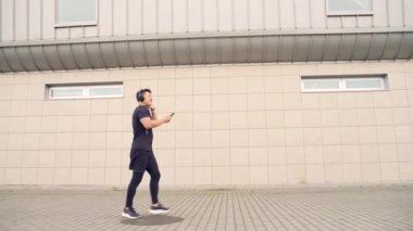 Genç, neşeli, aktif Asyalı, kulaklıkla gezen, müzik dinlemekten zevk alan, şehir merkezindeki modern sokak arka planında koşan sportif bir adam. Adam spor kulaklığı yapıyor.