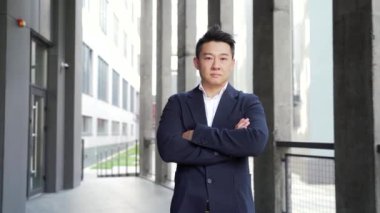 Genç özgüvenli, başarılı Asyalı iş adamı portresi kameraya bakıyor. Modern ofis binasının karanlık arka planında duran ciddi bir iş adamı. Erkek, kollarını kavuşturmuş resmi bir takım elbise.