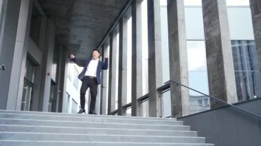 Neşeli mutlu Asyalı iş adamı dans ediyor merdivenlerden iniyor modern bir ofis binası, dışarıda, açık havada. Takım elbiseli komik başarılı iş adamı sevinçle atlıyor zaferi kutluyor, başarıyı.
