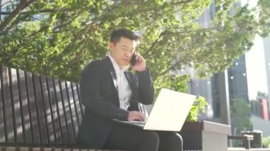 Genç Asyalı iş adamı, bankta oturan serbest çalışan, şehir merkezindeki modern kentsel sokak arka planında dizüstü bilgisayarla çalışan erkek, cep telefonuyla konuşan dış girişimcinin keyfini çıkarıyor.