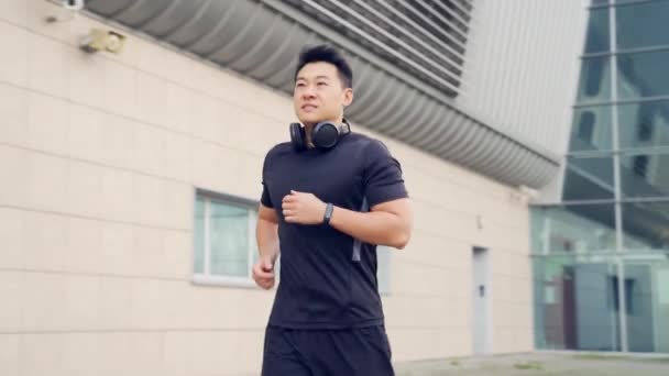 都会を背景に街を走り 公園でフィットネスウォッチのスマートブレスレットを楽しむ若いアジア人ランナー 黒のスポーツウェアで朝のジョギング アクティブな健康的なライフスタイルチェックトラッカー — ストック動画