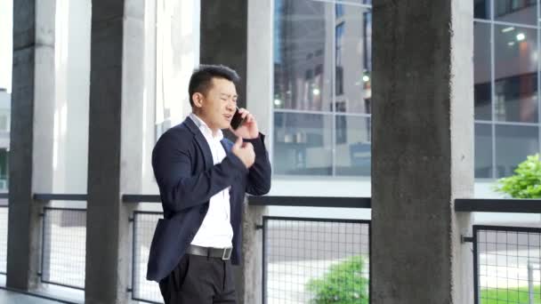 アジア系男性のビジネスマンは屋外で電話を持ってるビジネスマンの外の電話で怒ってる 緊張した狂牛病の専門家の会話スマートフォン 都市部の路上での刺激的な会話や歩行 — ストック動画