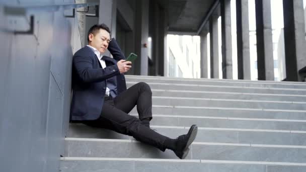 Młody Azjatycki Przygnębiony Człowiek Siedzi Schodach Zewnątrz Ulicy Pobliżu Biurowego — Wideo stockowe
