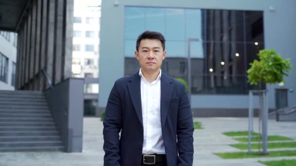 portrét mladého sebevědomého úspěšného asijského podnikatele hledícího do kamery. Vážný obchodník zblízka stojí na tmavém městském pozadí moderní kancelářské budovy venku. Muž ve formálním obleku