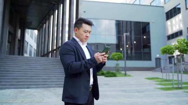 ダウンタウンのテキストメッセージ携帯電話の近代的なオフィスビルの都市の背景にアジアの従業員 ビジネスマンは スマートフォンの携帯電話を使用して正式なスーツやシャツのメッセージング立っている 屋外で — ストック動画