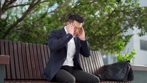 都会の公園のベンチに座っていた若いアジア系のサラリーマン 頭痛の種のマッサージの頭で過労の男性を強調した 苦痛に苦しむ疲れた従業員 病気の仕事 — ストック動画