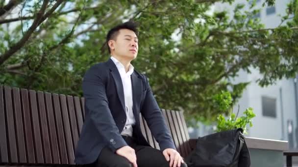 亚洲男性商人放松下来 坐在城市背景的长椅上 坐在城市公园的树木中间 在休息的时候 一位身穿正装的商人闭上了眼睛 — 图库视频影像