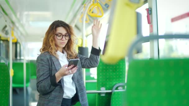 年轻的高加索女商人站在公共汽车内乘坐公共交通工具 女学生或女员工在无轨电车中使用手机 手持智能手机 站在现代有轨电车上 — 图库视频影像