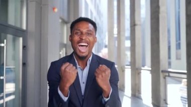 Heyecanlı Afrikalı Amerikalı iş adamı ofis binasının önünde kameraya bakarken zafere seviniyor. Resmi takım elbiseli mutlu serbest çalışan erkek, başarı, kazanma, kazanma ya da kazanma diye bağırıyor.