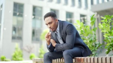 Afro-Amerikan iş adamı kenevir içiyor şehir parkındaki bankta oturuyor. Erkek işçi, iş adamı. Takım elbiseli bir ofis çalışanı dışarıda bir marihuanayı rahatlatıyor.