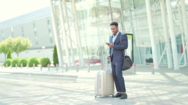 Arka planda duran Afro-Amerikan iş adamı resmi bir takım elbiseyle modern bir tren istasyonu havaalanında uygulamalı cep telefonu kullanan bir bavulla. Akıllı telefonlu gezgin bir iş adamı.