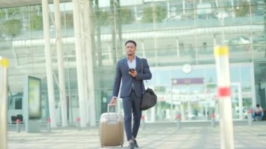 Afro-Amerikan turist işadamı, modern bir kentsel yapı sokağı olan terminal havaalanından bavulla geliyor. Elinde cep telefonu tutarak. Resmi takım elbiseli bir adam. seyahat