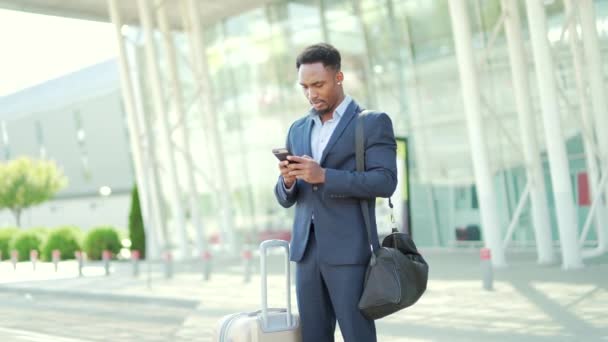 アフリカ系アメリカ人のビジネスマンが現代的な駅の空港を背景にアプリ携帯電話を使ったスーツケースを着て スマートフォンを手にビジネスマンの観光客を旅行 — ストック動画