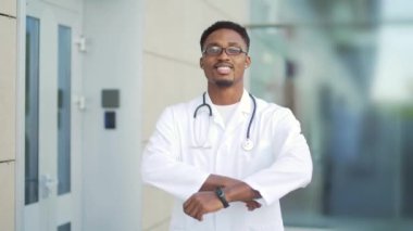 Afrika kökenli Amerikalı bir doktor kameraya bakıyor kollarını kavuşturup modern hastanenin arka planına bakıyor. Açık havada kendine güveni tam sağlıkçı. Erkek bilim adamı terapist kliniğin yakınında önlük giyiyor.